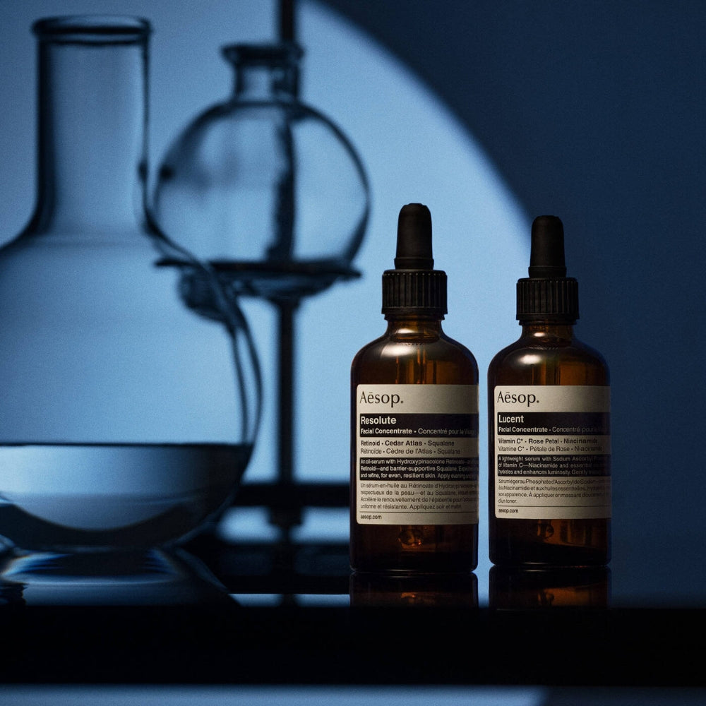 Two Aesop bottles with droppers on a reflective surface with blurred glassware in the background.