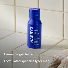 A blue bottle of Harry's Daily Facial Lotion SPF 15 stands on a beige tiled bathroom counter, with text below reading 'Dermatologist tested' and 'Formulated specifically for men'.
