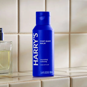 A blue bottle of Harry's Post-Shave Balm standing on a beige tiled surface, with part of a perfume bottle visible on the left.
